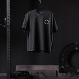 Black Tech Essential Relaxed Tee with circular logo, displayed on hanger in gym setting