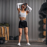 Black Lycra ribbed shorts worn with white sports bra and arm sleeves, model posing against corrugated metal backdrop